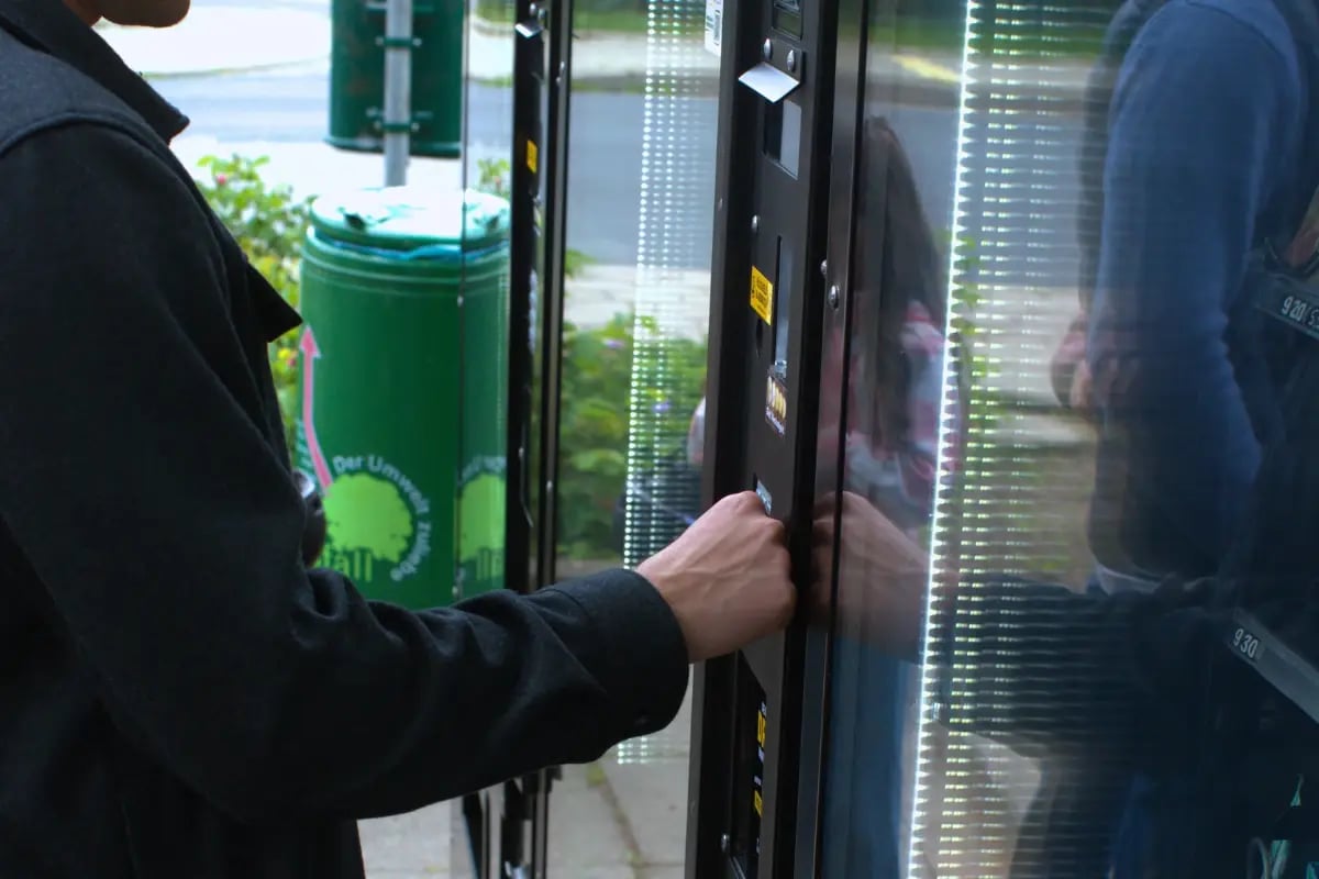 Bedienung eines Tante-Thea-Verkaufsautomaten: Kundin entnimmt ein Produkt aus dem Selbstbedienungsautomaten für die regionale 24/7-Nahversorgung in Hetlingen.